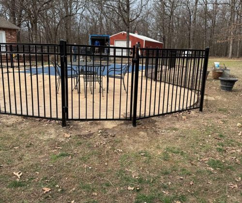 Black ornamental aluminum pool fence installation around concrete patio with gate access near wooded backyard in Northwest Arkansas