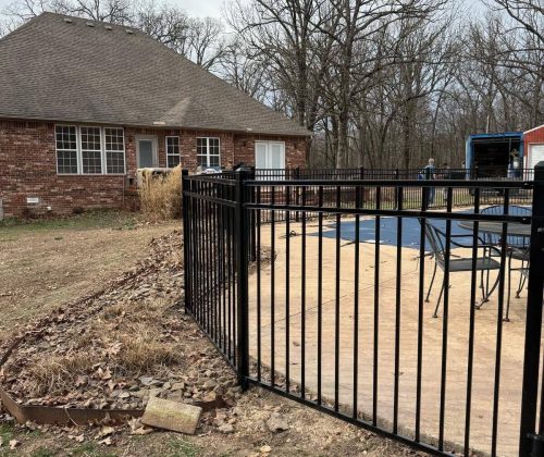 Black aluminum pool fence gate installation beside brick home with patio and winter landscaping in Northwest Arkansas