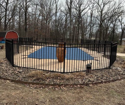 Black aluminum fence enclosing a covered backyard pool with stone edging and forest background in Northwest Arkansas