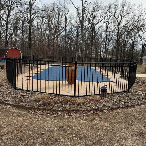 Black aluminum fence enclosing a covered backyard pool with stone edging and forest background in Northwest Arkansas