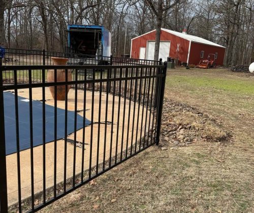 Black aluminum pool fence gate with covered pool and stone landscaping in a wooded backyard in Northwest Arkansas