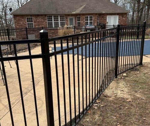 Wide view of black ornamental pool fence enclosing covered pool with curved layout and red shed in wooded Northwest Arkansas backyard