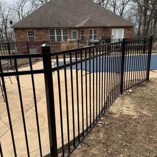 Wide view of black ornamental pool fence enclosing covered pool with curved layout and red shed in wooded Northwest Arkansas backyard